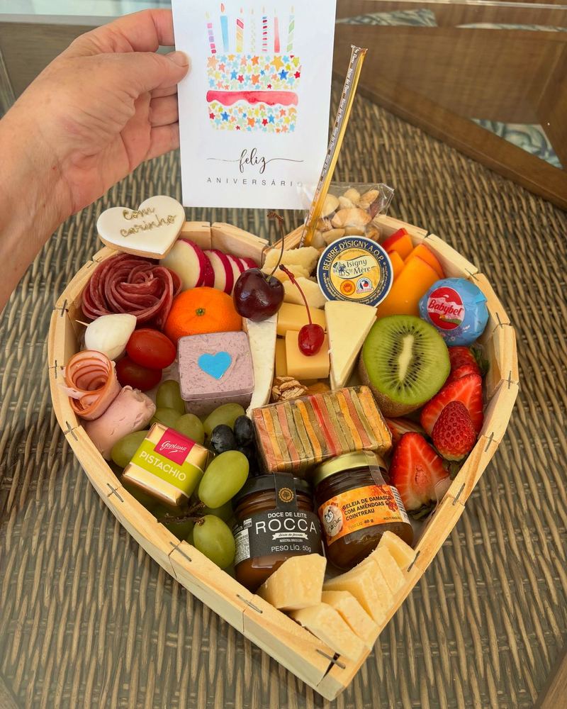 Caixa em formato de coração com frios, queijos e frutas, ao lado de um cartão de feliz aniversário